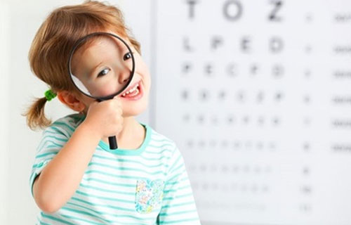 Ein Kind sitzt in der Ordination bei einem Augenarzt und hält sich eine Lupen vor das Auge